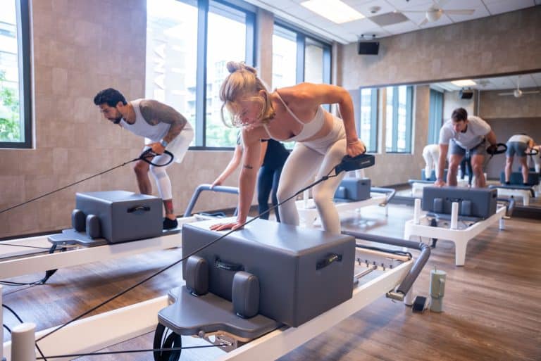 People exercising on reformer machines in a well-lit fitness studio with large windows and wooden flooring.