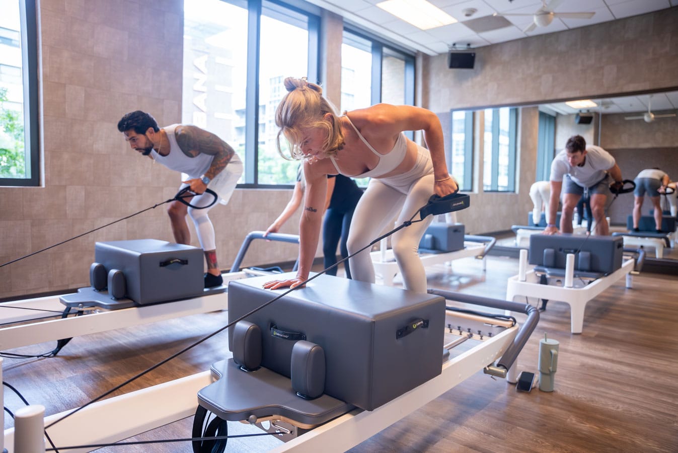 People exercising on reformer machines in a well-lit fitness studio with large windows and wooden flooring.