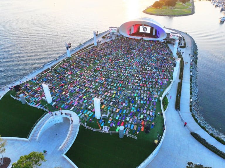 A large outdoor event at a waterside amphitheater with a crowd seated on colorful mats, facing a stage with a white, domed roof at sunset.