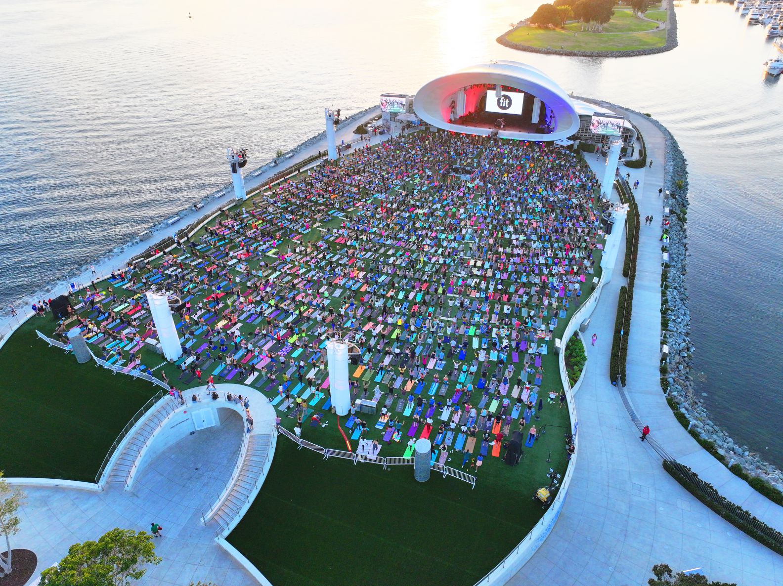 A large outdoor event at a waterside amphitheater with a crowd seated on colorful mats, facing a stage with a white, domed roof at sunset.