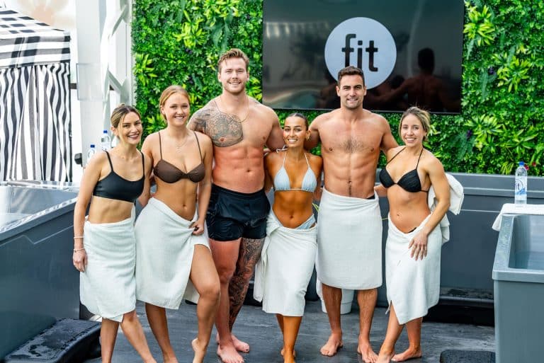 A group of six people wearing swimwear and wrapped in towels stand together smiling in front of a "fit" sign and a green wall.