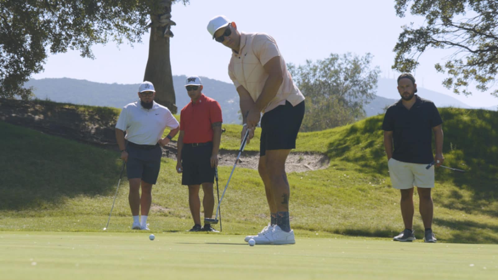 Four men playing golf on a sunny day. One man is putting on the green while the others watch. Trees and hills are in the background.